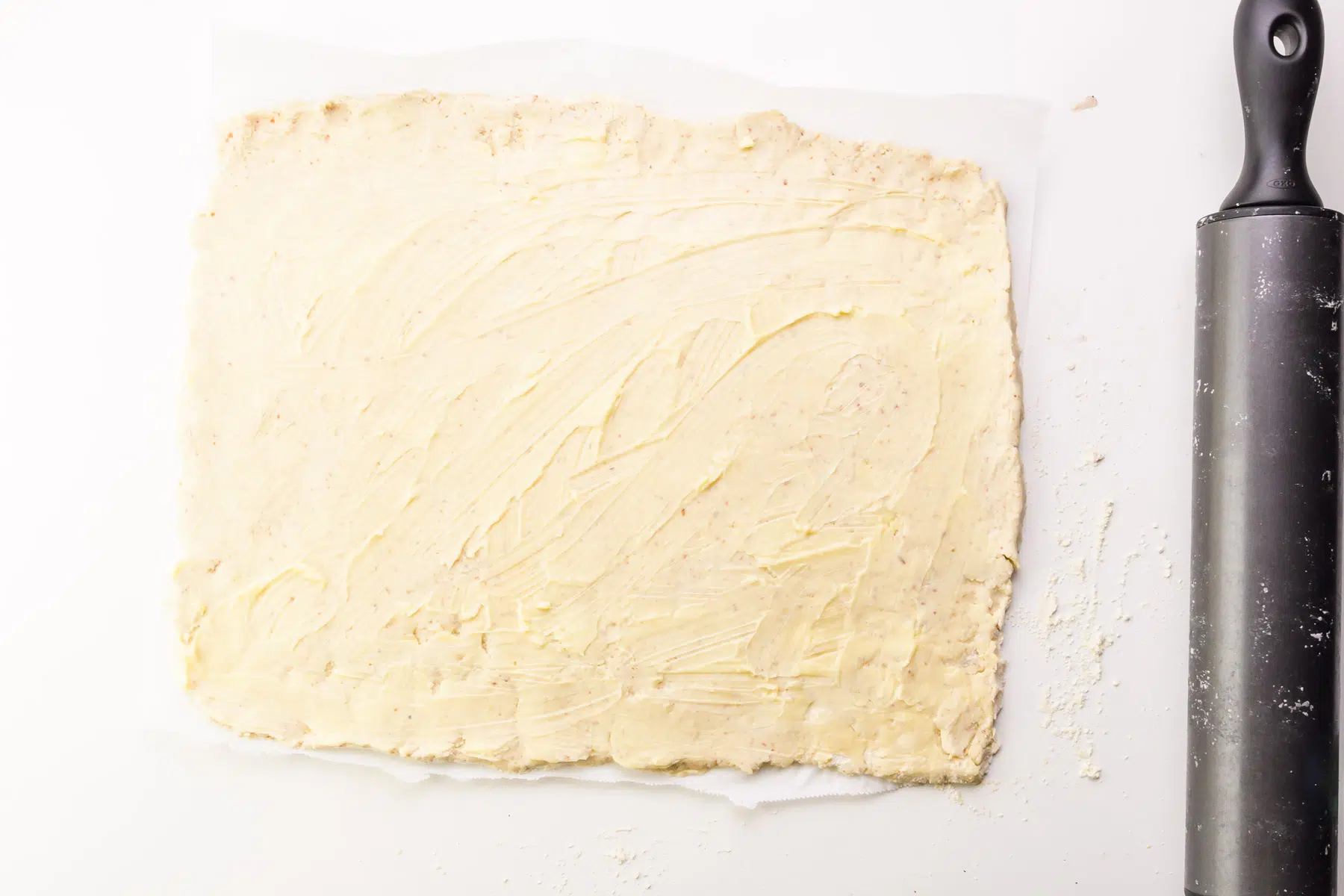 Rolled out dough has a layer of vegan butter spread on top. It sits beside a rolling pin.
