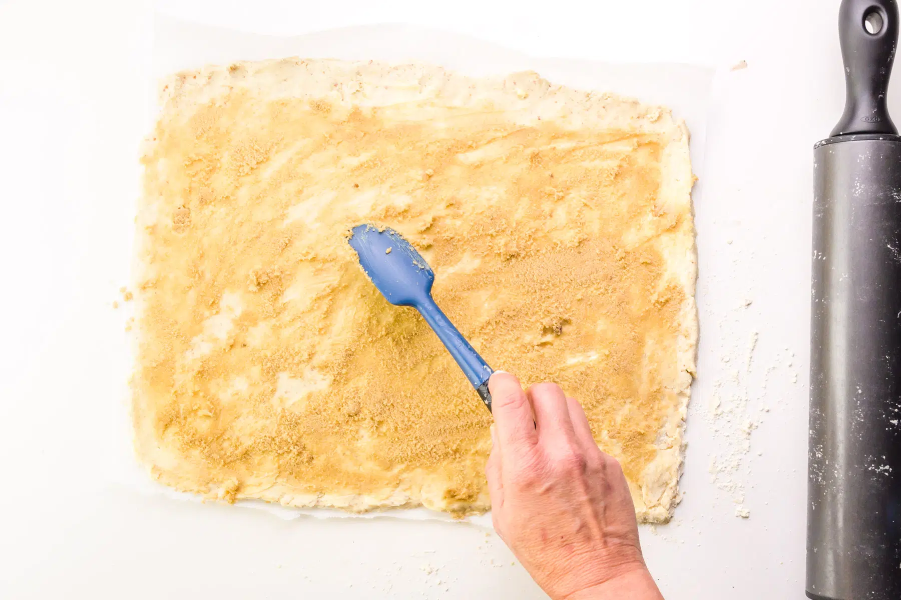 A hand holds a spatula spreading brown sugar over rolled out dough. There's a rolling pin on the right side.