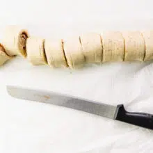 A serrated knife sits below a log of dough that has been sliced into a number of rolls.