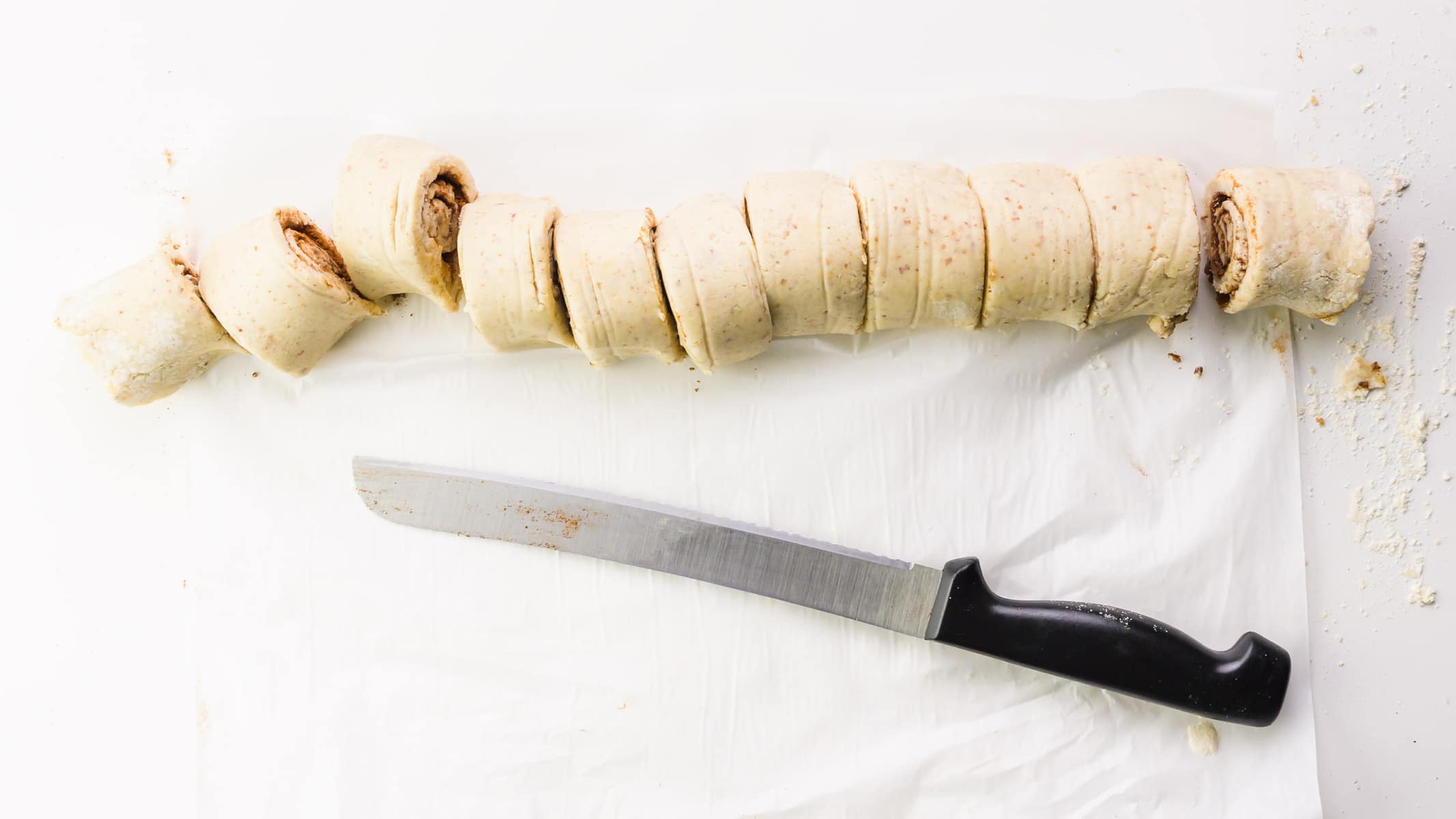 A serrated knife sits below a log of dough that has been sliced into a number of rolls.