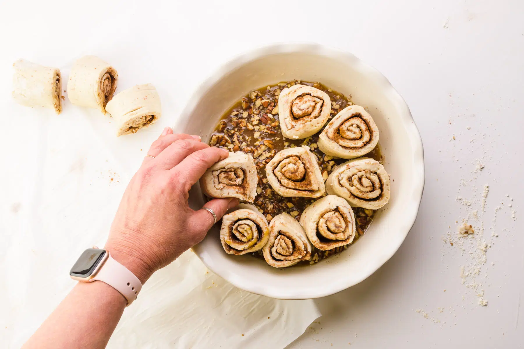 A hand places a cinnamon roll in a white pan with more rolls. There are more cut rolls on the left.