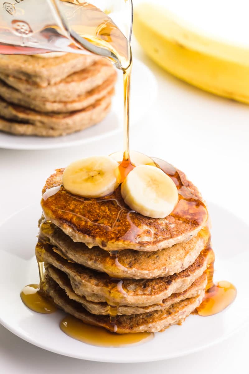 Syrup is being poured from a pitcher over a stack of pancakes with sliced bananas on the top. There's another stack of pancakes next to a banana in the background.