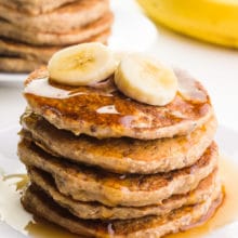 A plate holds several gluten-free banana pancakes with syrup on top. There are sliced bananas on top of the stack. There's another stack of pancakes on a plate next to a banana in the background.
