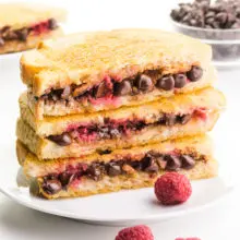 A stack of four chocolate grilled sandwiches are on a plate and shows melted chocolate and raspberries. There are fresh raspberries around the plate and another sandwich and bowl of chocolate chips in the background.