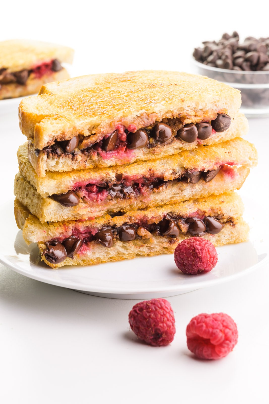 A stack of grilled sandwiches sits on a plate. They showcase melted chocolate and raspberries. There are fresh raspberries around the plate, and another sandwich and a bowl of chocolate chips in the background.