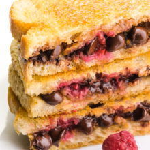 A stack of three chocolate grilled sandwiches shows melted chocolate chips and raspberries with a golden crust on the top slice of bread. There is a raspberry sitting in front of the stack.