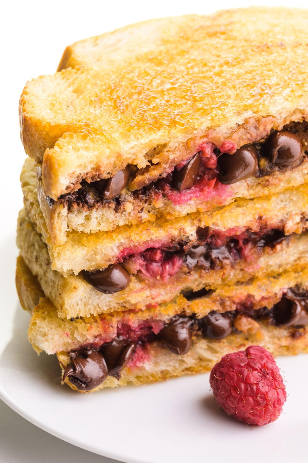A stack of three chocolate grilled sandwiches shows melted chocolate chips and raspberries with a golden crust on the top slice of bread. There is a raspberry sitting in front of the stack.