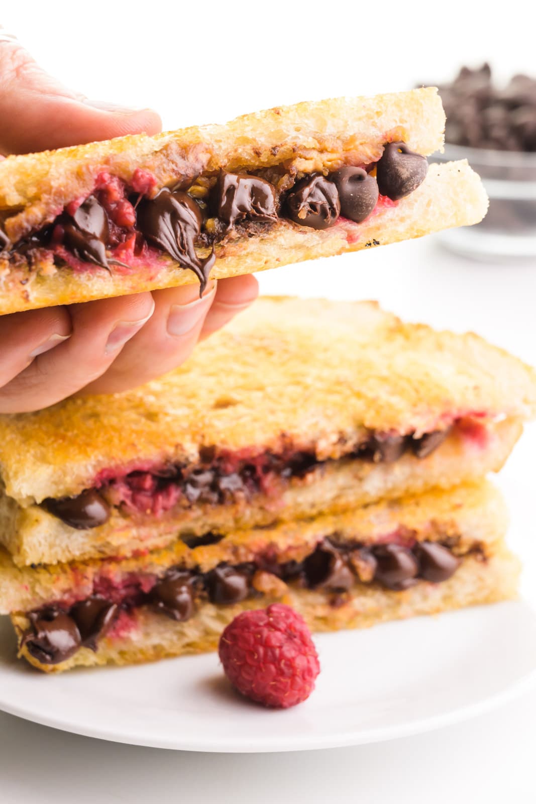 A hand holds a grilled chocolate sandwich hovering over a sack of two more of the sandwiches on a plate. There is a fresh raspberry on the plate and a bowl of chocolate chips in the background.
