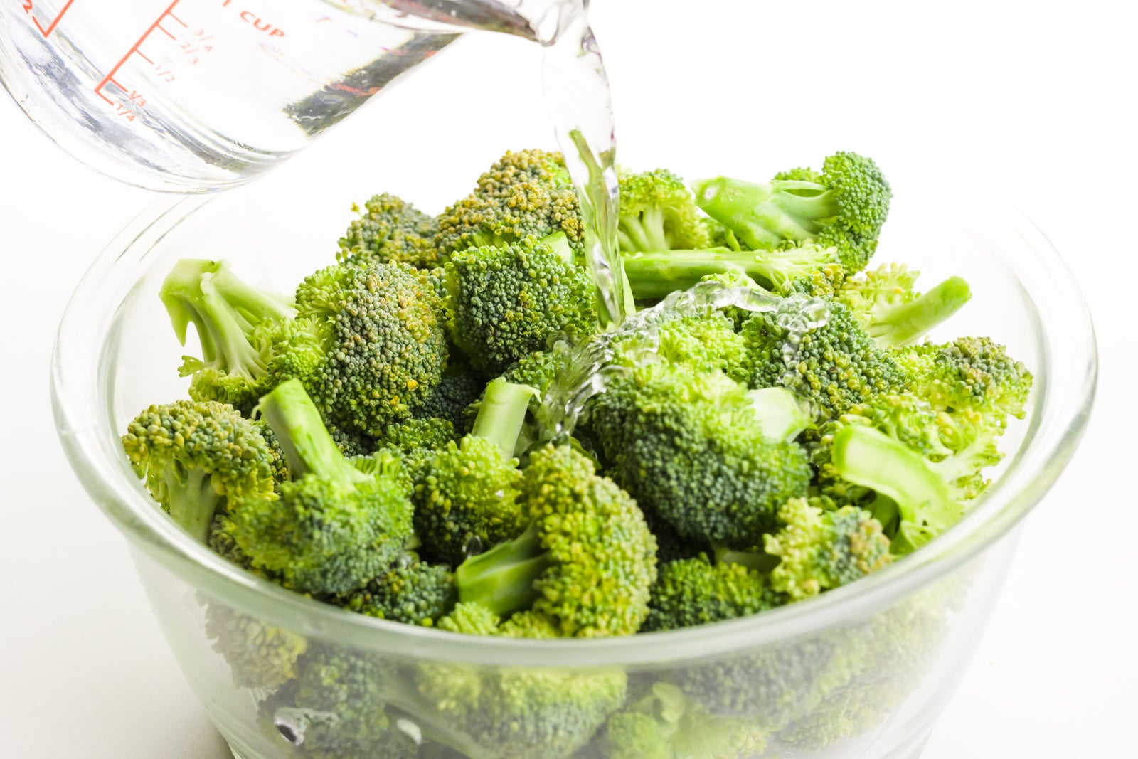 A pyrex measuring cup is full of water that is being poured over a bowl of broccoli florets.