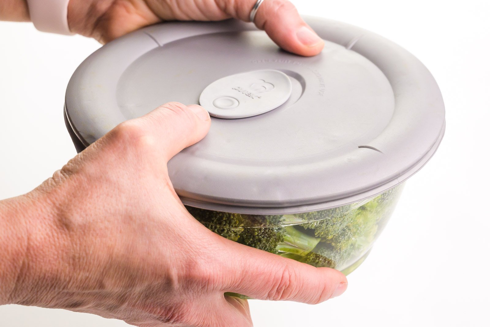 Two hands hold a lid over a bowl of broccoli florets and water. The hands are shaking the bowl to wash the florets.