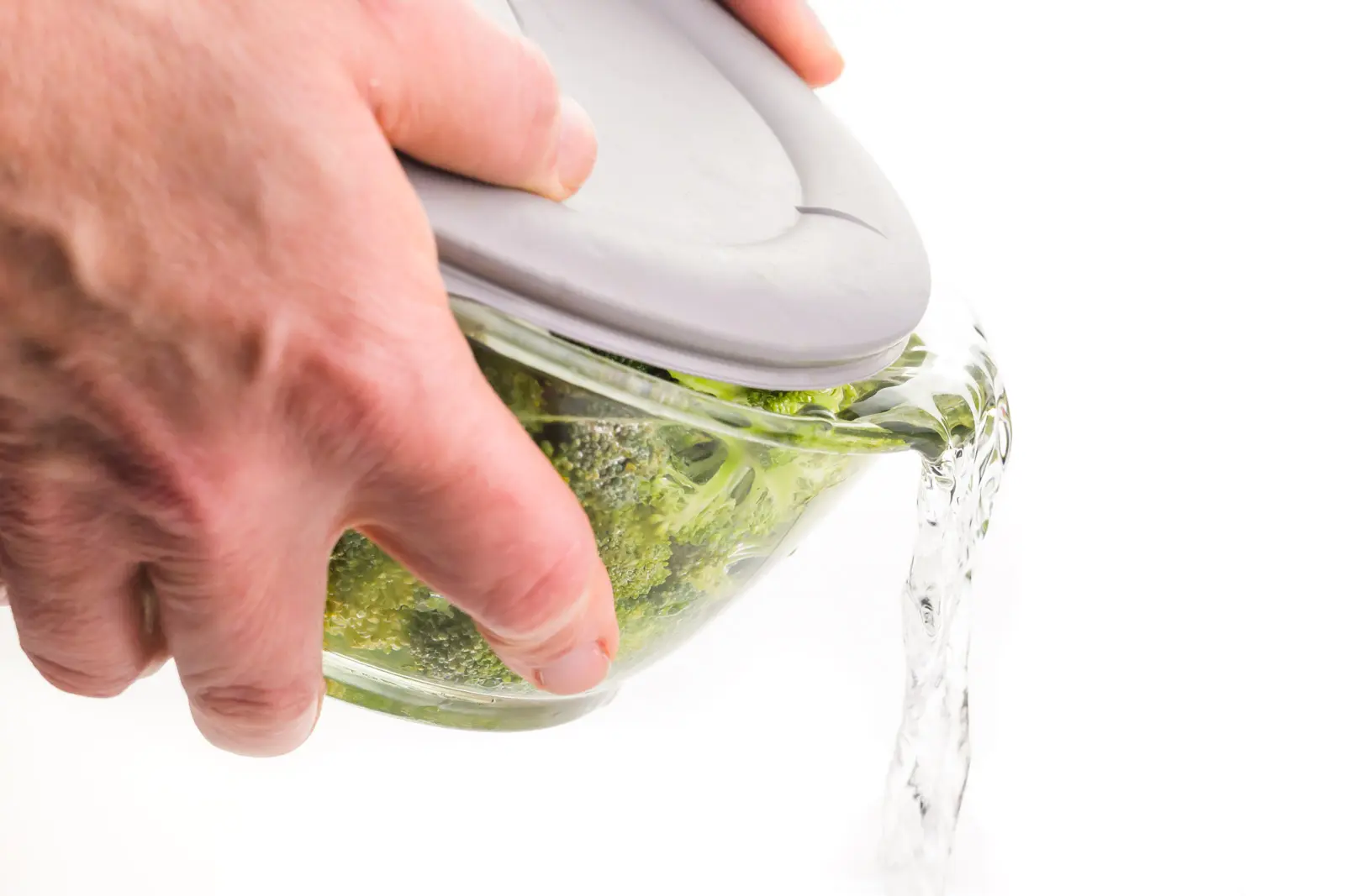 Two hands hold a lidded bowl full of broccoli florets and water. The lid is slightly ajar and the water is being poured out of the bowl.