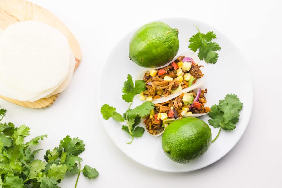 Looking down on two jicama tacos on a plate with limes and fresh cilantro. There is more fresh cilantro and jicama tacos on the left.