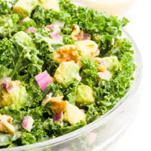 A large bowl is full of raw kale salad, showing bits of avocado and walnuts on top. There is more dressing and a chopped red onion in the background.