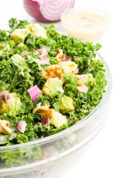 A large bowl is full of raw kale salad, showing bits of avocado and walnuts on top. There is more dressing and a chopped red onion in the background.