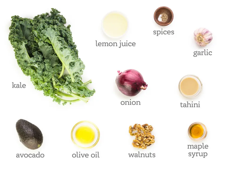 Ingredients are on a white table. The labels next to them read, Kale, lemon juice, spices, garlic, tahini, maple syrup, walnuts, onion, olive oil, and avocado.