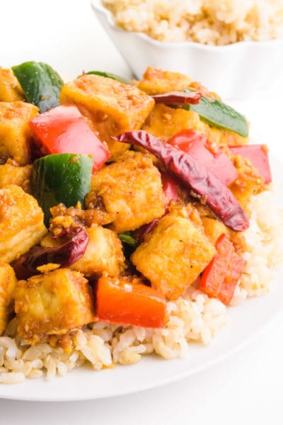 A dish of kung pao tofu is served over rice. There is a bowl with more rice in the background.