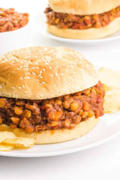 Two buns sit on plates. There is a lentil sloppy Joe mixture in the buns. There are potato chips next to the front plate and a bowl with more lentil mixture by the back plate.