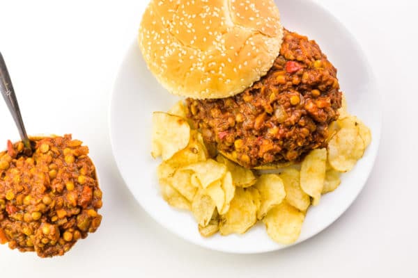 Looking down on a plate with a vegan sloppy Joe mixture on a bun, with the top bun to the side. There are potato chips on the plate, too. Next to the plate is a bowl with more of the vegan mixture with a spoon in in it.