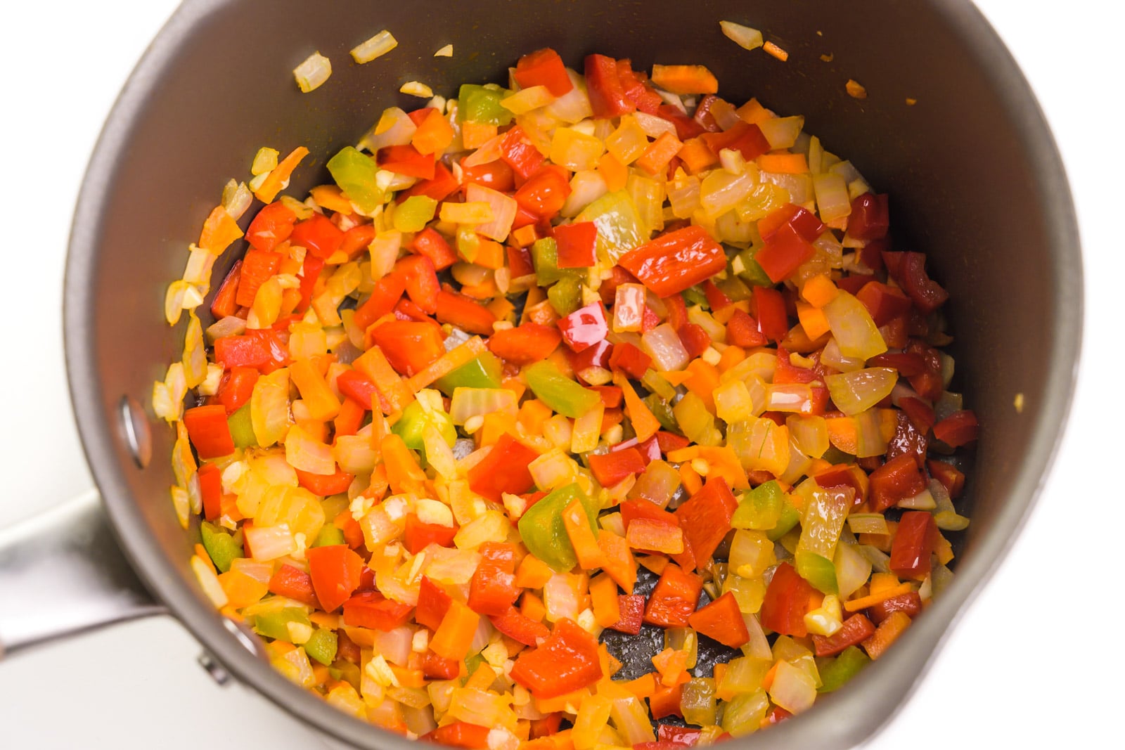 Looking into a saucepan with chopped veggies, such as onions and red bell peppers, being cooked until tender.