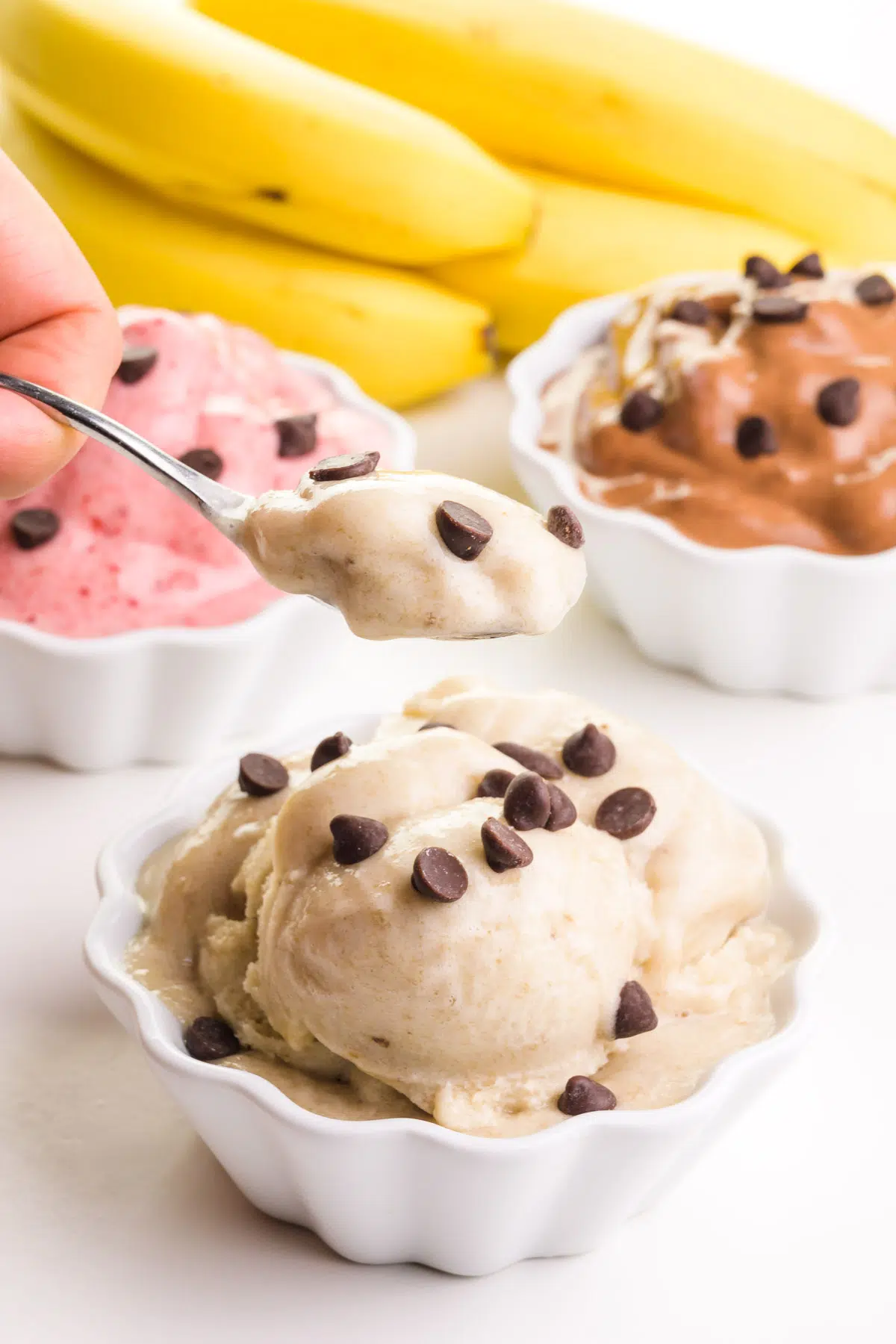 A hand holds a spoonful of banana ice cream hovering over a bowl of it. There are different flavors of ice cream and bananas in the background.