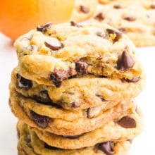 A stack of cookies shows the top one with a bite taken out, revealing melted chocolate chips. There are more cookies and an orange in the background.
