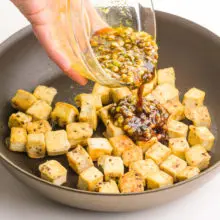 A hand holds a bowl of sauce and is pouring it into a skillet with tofu cubes.