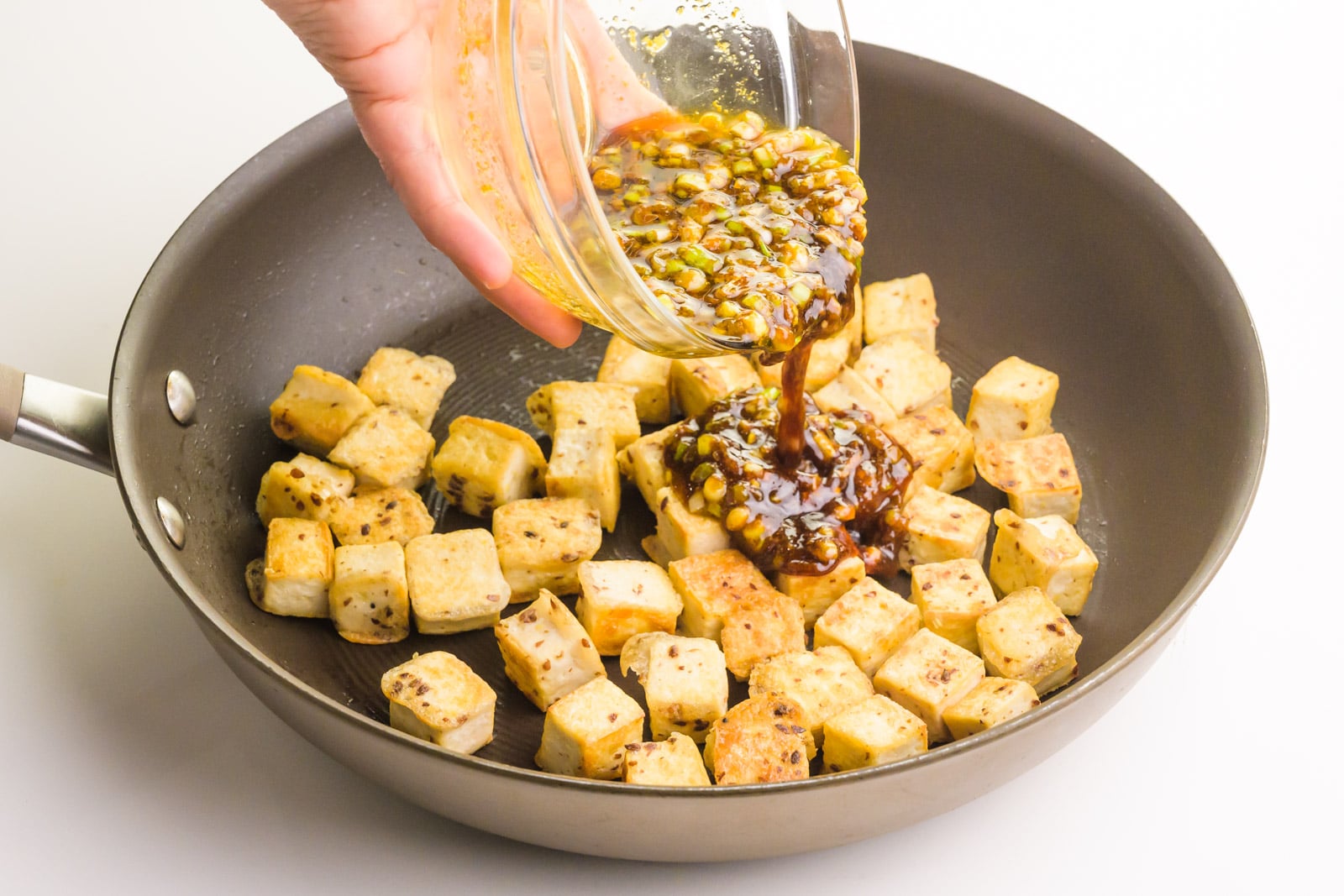 A hand holds a bowl of sauce and is pouring it into a skillet with tofu cubes.