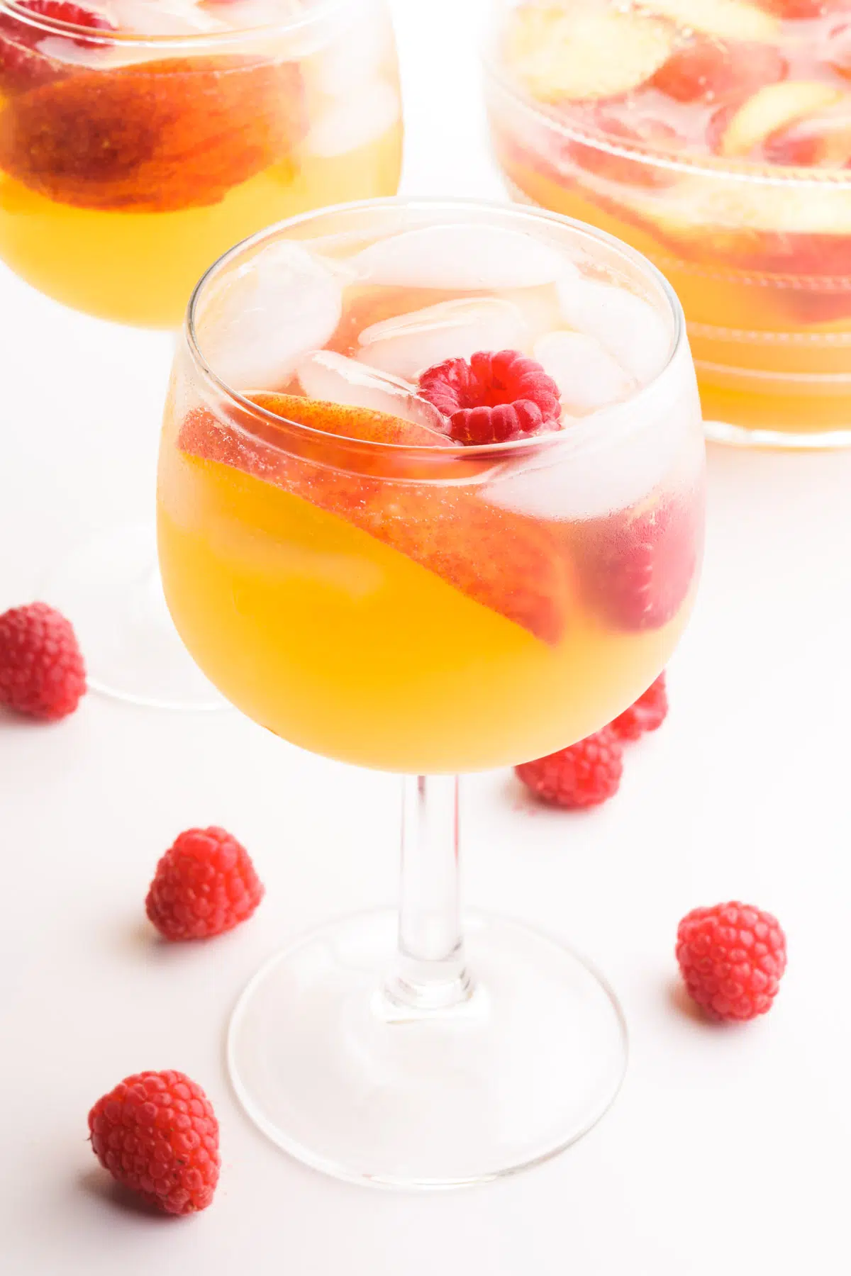 A wine glass holds peach sangria with fresh raspberries and peach slices. There's fresh raspberries, a pitcher of sangria, and another wine glass full of sangria around it.