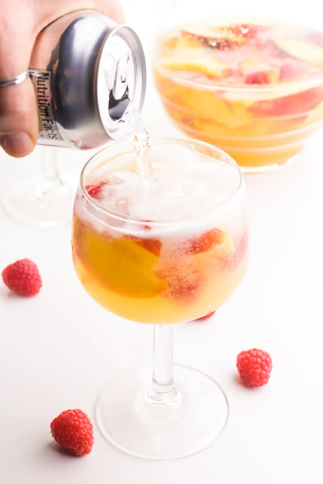 A hand holds a can of club soda and is pouring it into a wine glass full of sangria. There are fresh raspberries around the glass and a pitcher of sangria in the background.