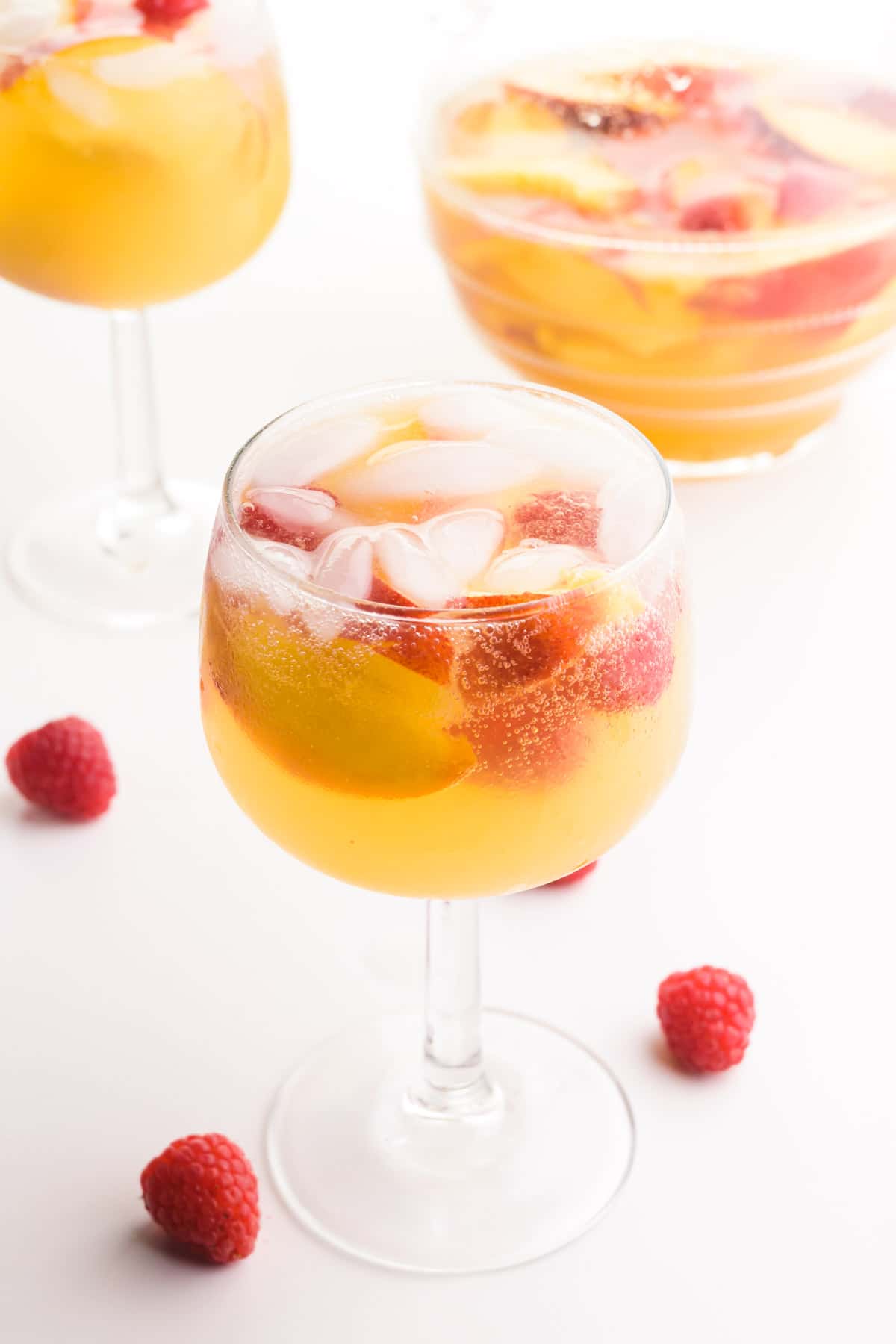 White peach sangria is in a wine glass along with peach slices and fresh raspberries. There are fresh raspberries, another glass of sangria and a pitcher of sangria in the background.