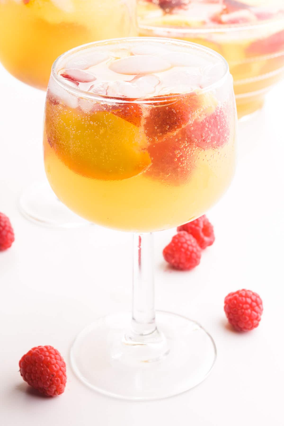 A wine glass holds peach sangria, ice cubes, raspberries, and peach slices. There is another glass and a pitcher in the background. There are fresh raspberries on the table beside the glasses.