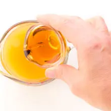 A hand holds a bowl with maple syrup and is pouring it into a glass measuring cup with peach juice in it.