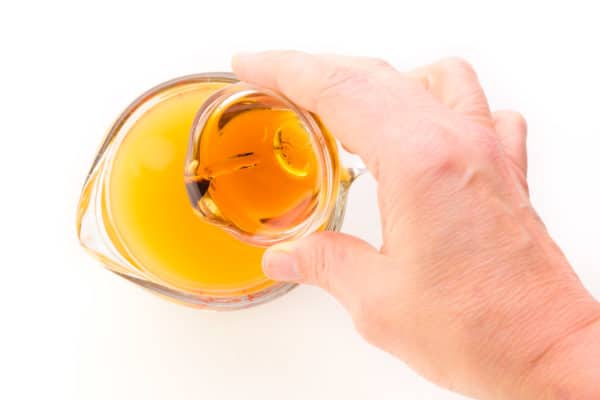 A hand holds a bowl with maple syrup and is pouring it into a glass measuring cup with peach juice in it.