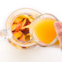 A hand holds a glass measuring cup full of peach juice and is pouring it into a pitcher with sliced peaches.