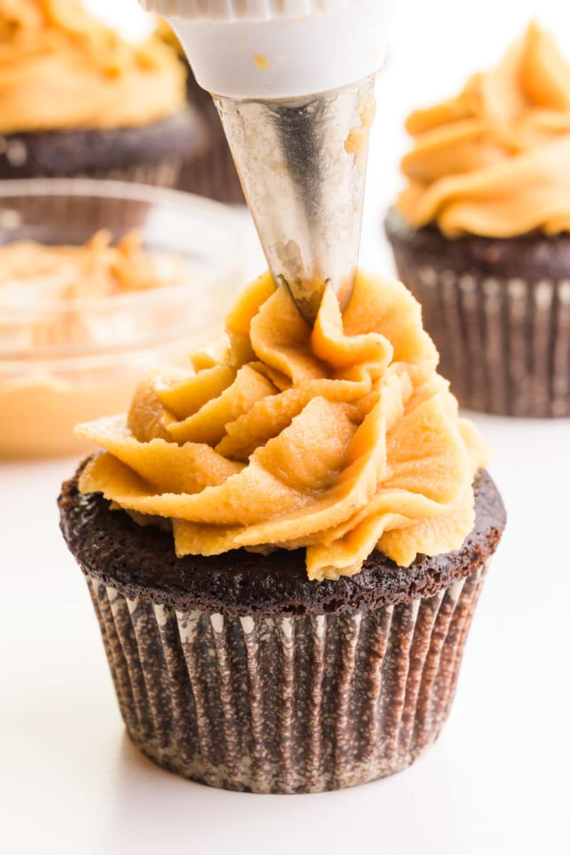 Peanut butter frosting is being piped in a fancy swirl on top of a chocolate cupcake. There's a bowl of peanut butter and more frosted cupcakes in the background.