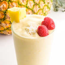 A glass of pineapple smoothie has fresh raspberries, coconut flakes, and a pineapple chunk on top. It sits in front of a whole pineapple.