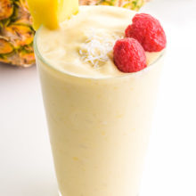 A glass full of Pina colada smoothie has raspberries, coconut flakes, and a pineapple chunk on top. It sits in front of a pineapple.