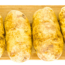 Four potatoes have been picked with a fork, covered with oil, and seasoned with salt and pepper. They are sitting on a cutting board.