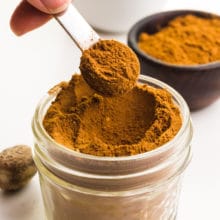 A hand holds a teaspoon of pumpkin pie spice hovering over a glass jar full if it. There are more spices in bowls around it.