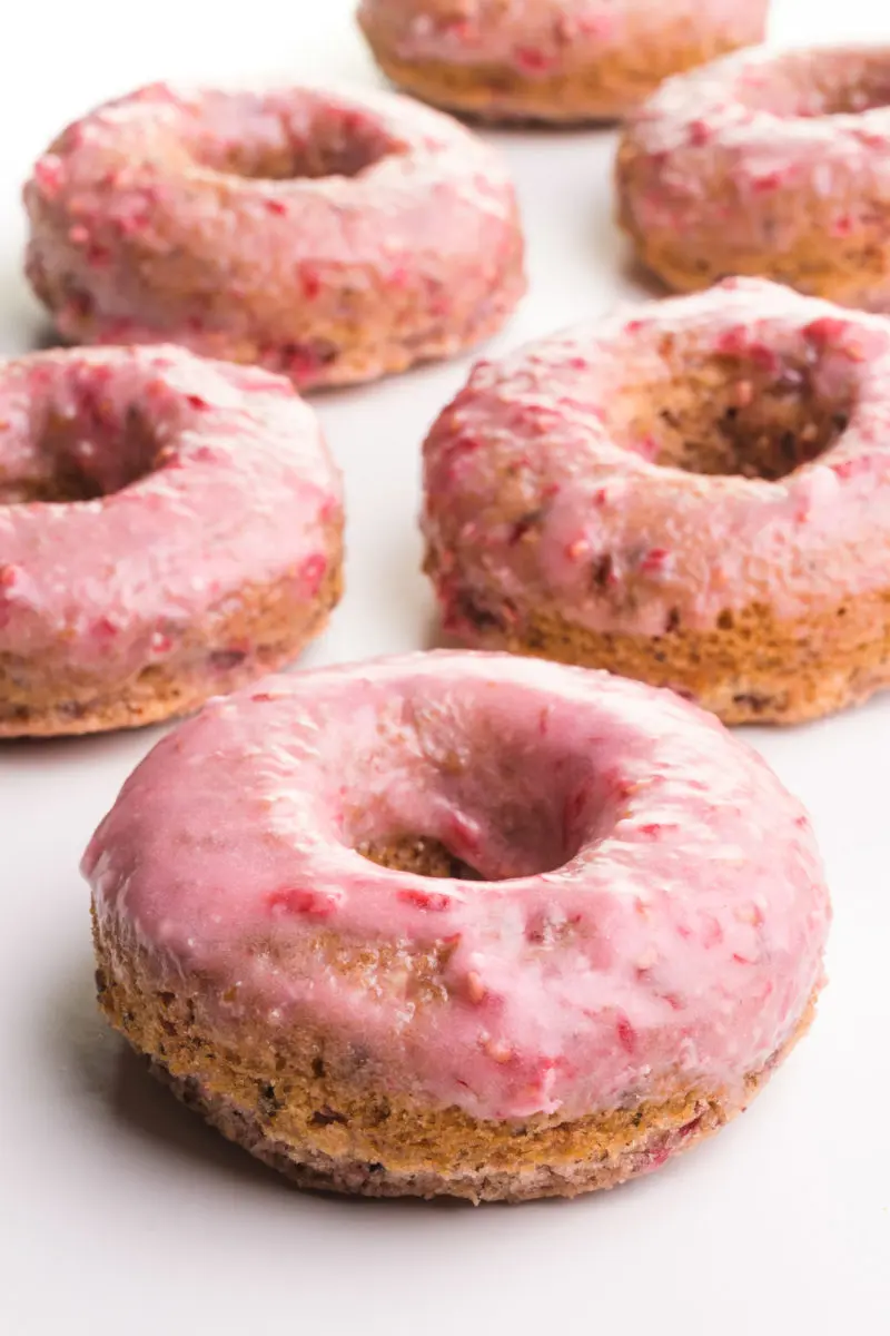 A raspberry donut has pink frosting on top and is surrounded by more donuts in the background.