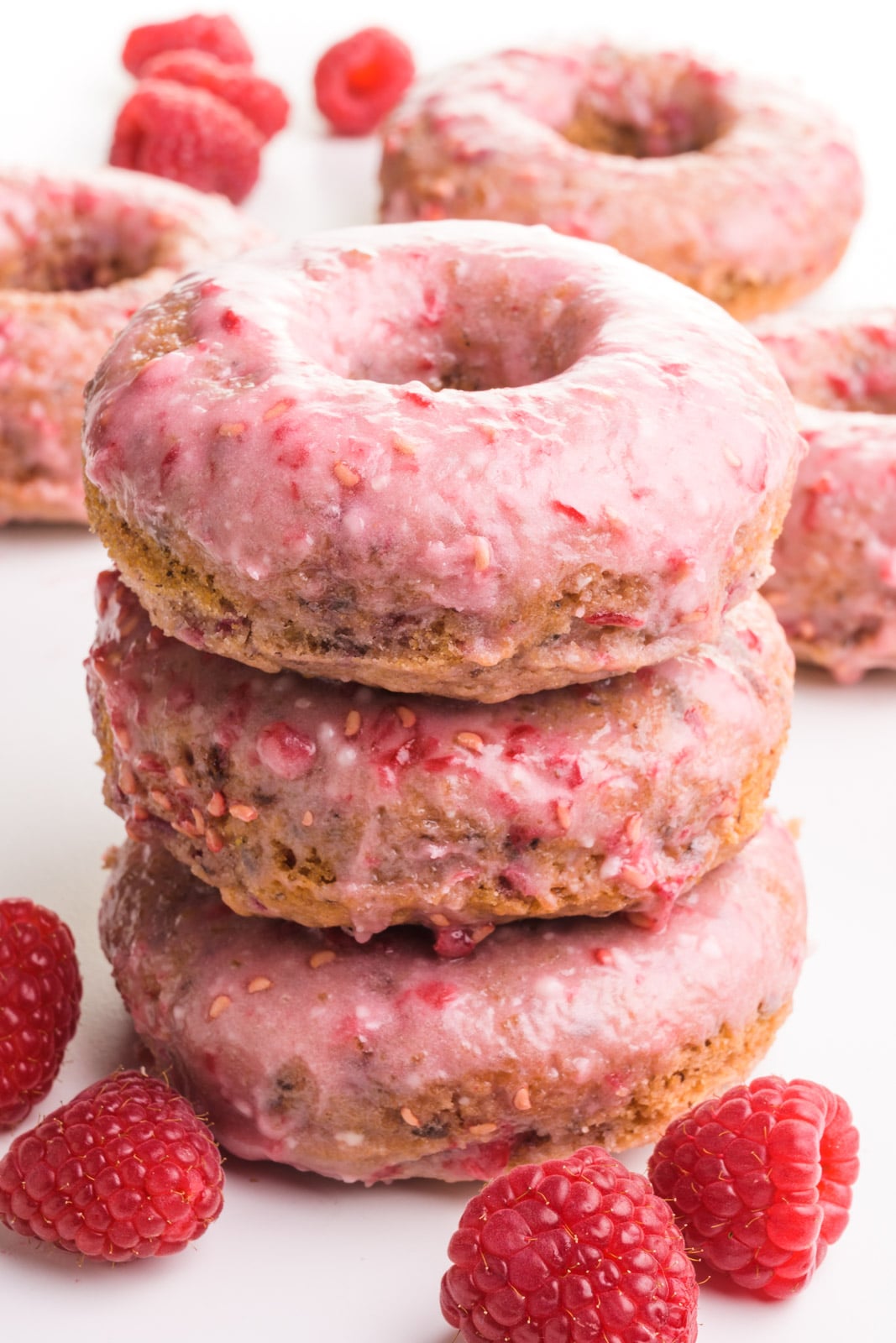 A stack of three donuts has fresh raspberries and more donuts around it and in the background.