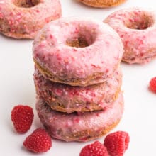A stack of three pink donuts are surrounded by fresh raspberries. There are more donuts in the background.