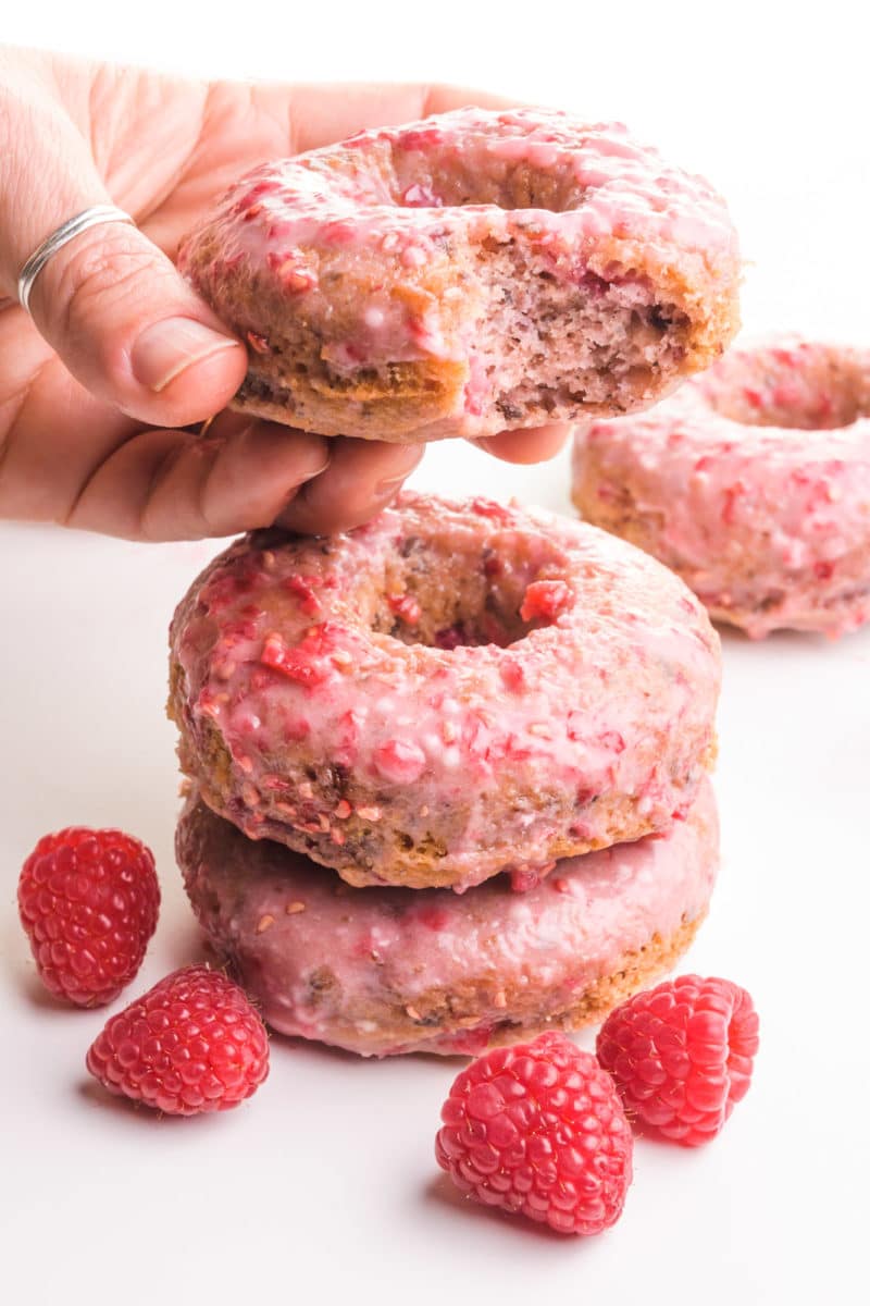 A hand holds a raspberry donut with a bite taken out. It's hovering over a stack of two more donuts with fresh raspberries around it. There's another donut in the background.