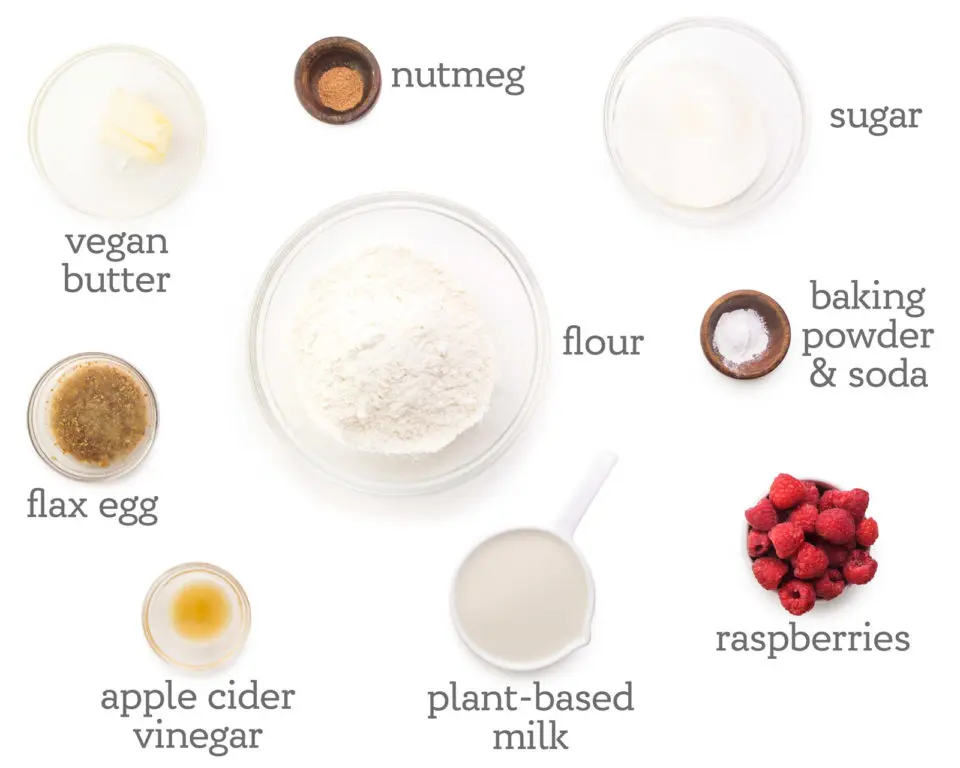 Ingredients are laid out on a white counter. The labels next to them read, sugar, baking powder & soda, raspberries, plant-based milk, apple cider vinegar, flax egg, vegan butter, nutmeg, and flour.