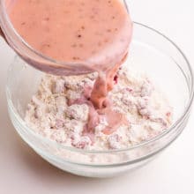 A hand holds a bowl pouring a pink milk mixture into a bowl with flour.