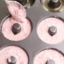A spoon is drizzling pink batter into donut pan compartments.