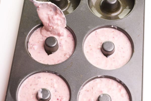 A spoon is drizzling pink batter into donut pan compartments.