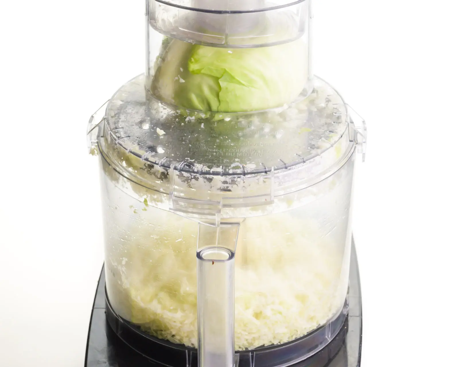 Cabbage is in the top of a food processor, using the shredding blade to shred it. There is shredded cabbage in the main bowl of the food processor.
