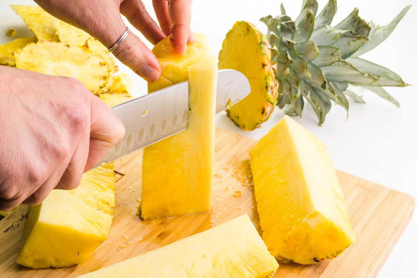 A hand holds a knife cutting down on a quartered piece of a pineapple, removing the core. There are pieces of pineapple sitting next to it on a cutting board.