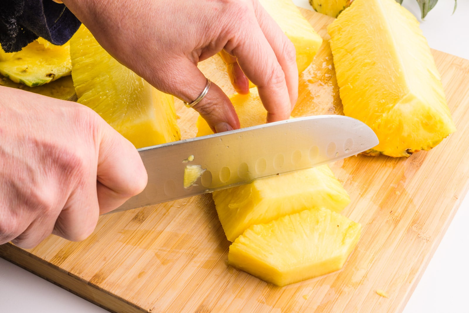 A hand holds a knife cutting skinned pineapple strips into chunks. There are pieces of the pineapple sitting around it on a cutting board.